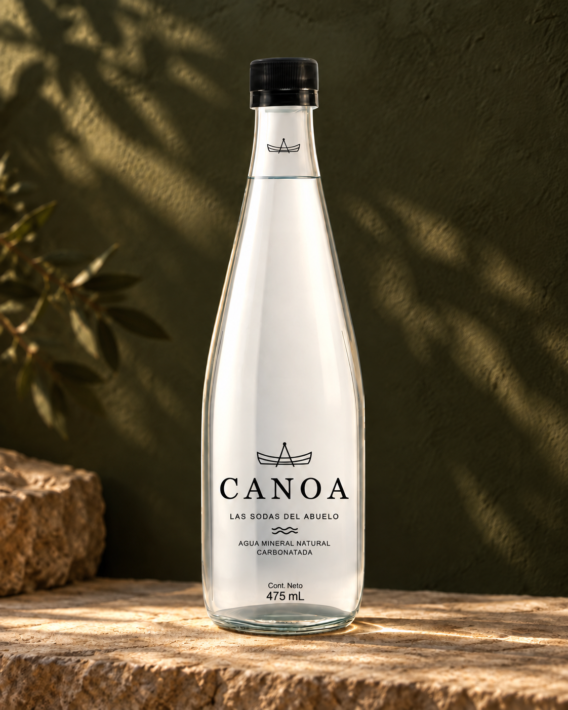 Botella CANOA de 475 ml con la etiqueta Agua mineral natural carbonatada, letras negras y tapa negra, sobre piedra y frente a un fondo verde oliva con sombras de hojas.